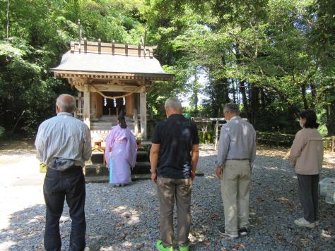 小さな神社の前に神職の人が立ちその後ろに頭を下げた男女が立っている写真