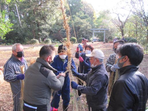 鳥居が奥に見える雑木林の開けた場所で数名の男女が藁をもって作業している様子の写真