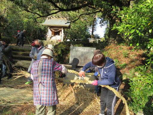 石碑の前で数名の男女が編まれた藁を持ち作業を行う様子の写真