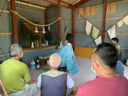 トタン小屋内部の本尊前で和装の男性が祈りを捧げその周りを男性数名が囲んでいる写真