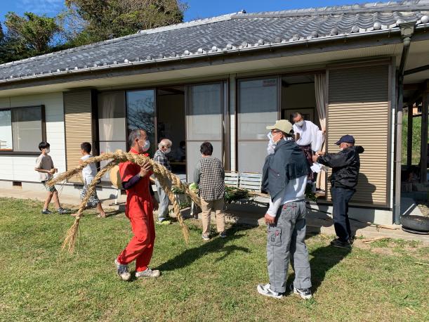 複数の男女が家の前の庭で晴れの日に縄やお供え物を持って準備している写真