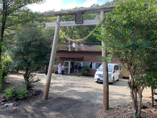 しめ縄がついた鳥居の後ろに4人の人がいて建物と車が置いてある写真