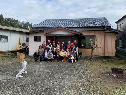 瓦屋根の一軒家の前でカメラマンの後ろから撮影された50周年記念の老若男女の集合写真