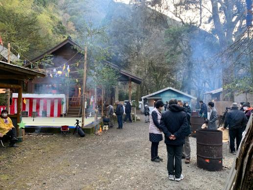 森に囲まれた神社の前で複数の男女がストーブのようなもので暖を取っている写真