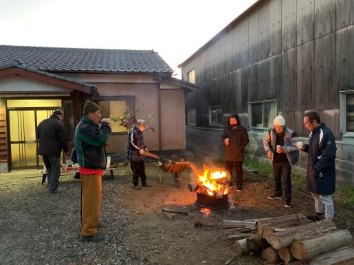 平屋の前で厚着をした男性数名が焚火を囲んで暖を取っている写真