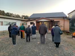 日の出の時刻に合わせ平屋の前で奥を向いて手を合わせ祈っている人たちの写真