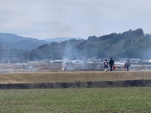 曇りの日に人が歩いているあぜ道と山の間にある田畑から煙が上がっている様子の写真