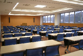 横長の机が数列並べられていて、それぞれに青い椅子が三脚ずつ設置されている室内の写真