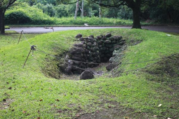 草が周りに生えており真ん中がくぼんでいる土地に黒い石が積み上げられている古墳の写真