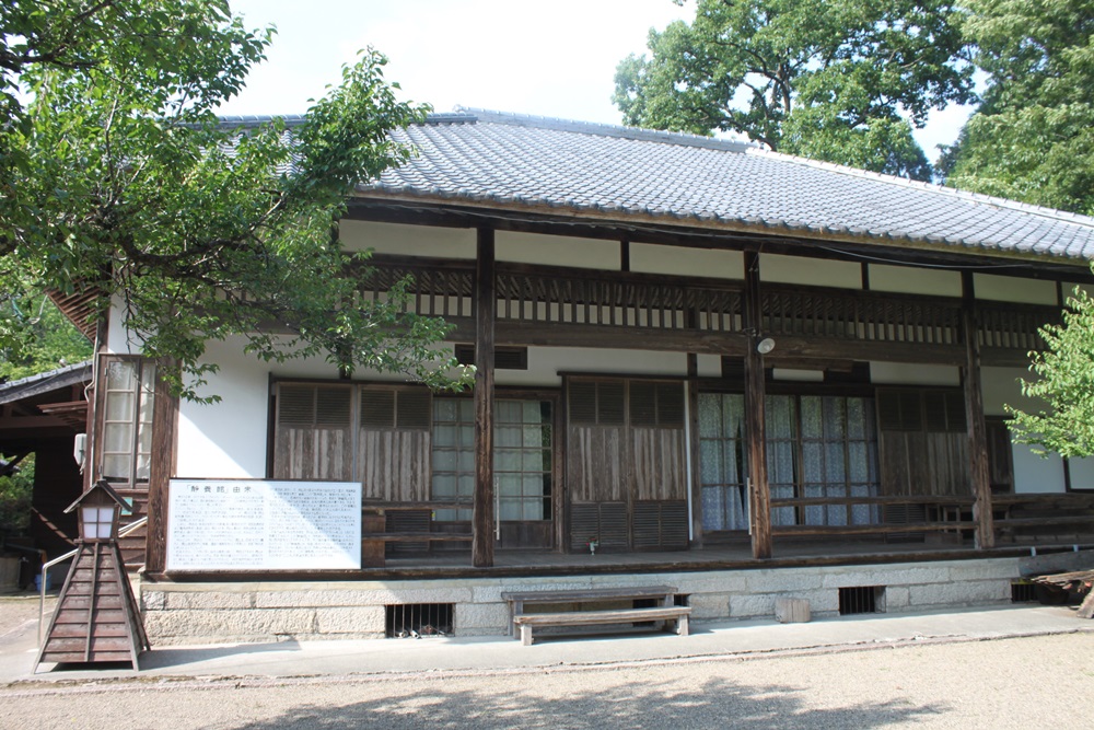 周りに木が生い茂っている真ん中に瓦屋根の平屋建ての建物と手前に行灯がある建物の写真